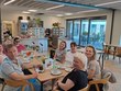 Na fotografii je zachycena skupina lidí. Zleva: p. Limberk, p. Parkosová, DiS., p. Oherová, p. Bohuslavová, p. Špinarová, DiS., p. Neckářová, ředitelka p. Mgr. Bělovová Dolejší, p. Limberková. Všichni sedí u společného stolu v útulné kavárně. Na stole jsou šálky s kávou, nápoje a drobné občerstvení. Usmívají se a působí velmi příjemně a uvolněně. V pozadí je kavárenský pult s vitrínami a za skleněnými dveřmi venkovní posezení