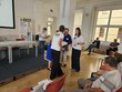 Na fotografii probíhá soutěž v Braillově písmu. V místnosti se pohybují účastníci a organizátoři, kteří spolu komunikují. Několik soutěžících sedí na židlích a čeká, další lidé stojí uprostřed místnosti. U stěny je stůl a informační plakát, místnost je světlá a prostorná